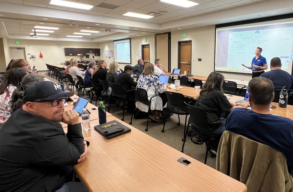 People sitting in a classroom setting listen to a presenter near a projection screen displaying a presentation. Everyone has laptops, and the room is well-lit with a modern conference design.