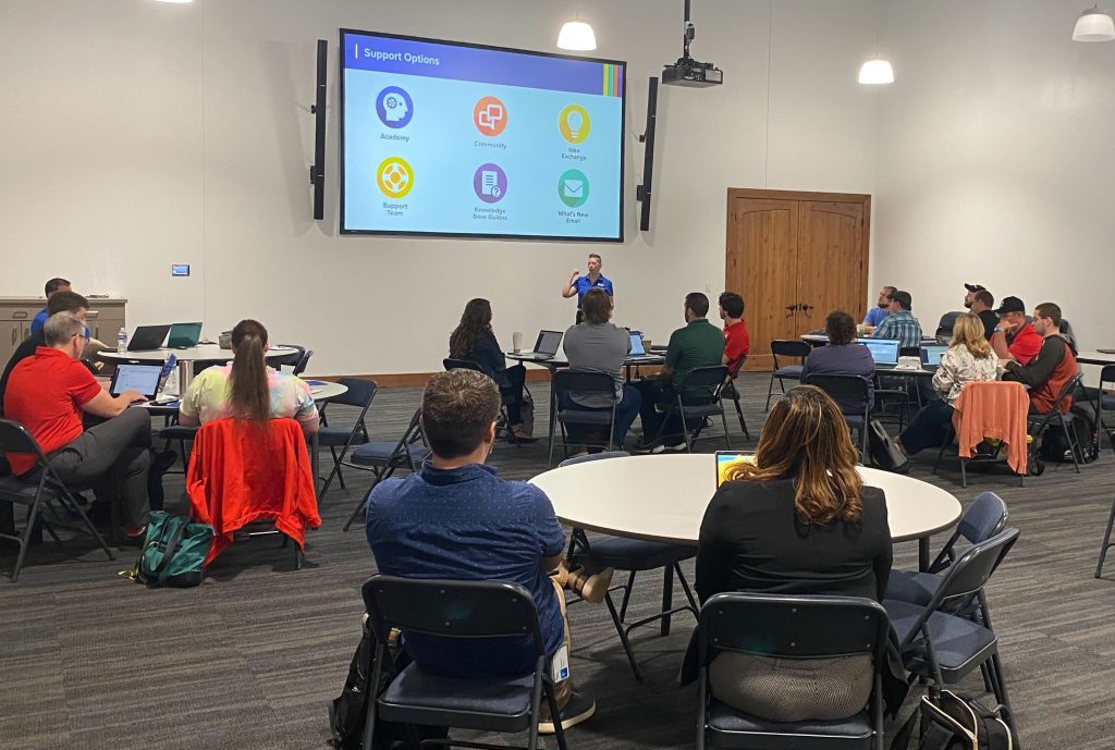 A person presents in a conference room, gesturing towards a screen displaying "Support Options" with icons like Academy, Community, and Support Team, while attendees sit in circular tables, observing.