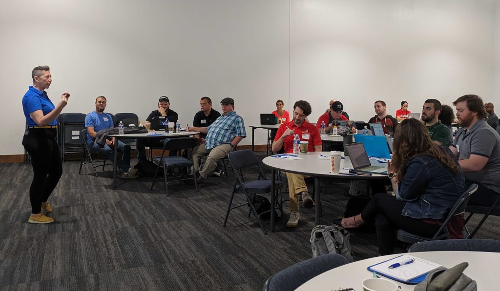 A speaker stands gesturing, addressing a group of people seated around tables with laptops and notebooks, in a conference room with a plain wall.